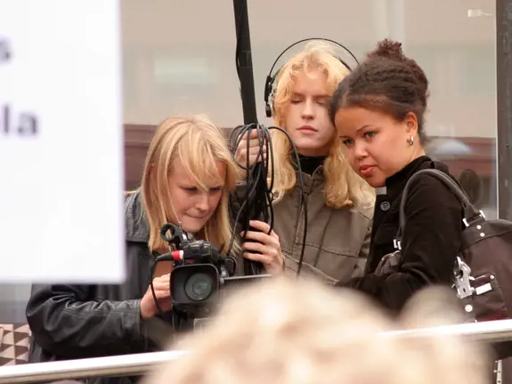 Tre unga kvinnor står utomhus och håller på att filma. Kvinnan längst till vänster håller i en kamera, kvinnan i mitten har hörlurar och håller i sladdar till kameran. Kvinnan som står till höger tittar koncentrerat mot det de filmar.