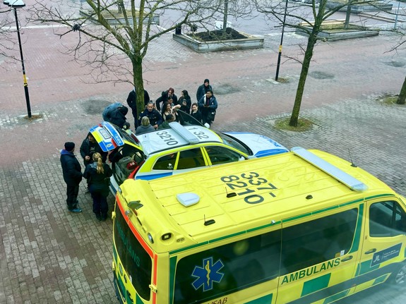 Deltagare från Röda Korsets folkhögskola står samlade utanför skolan vid en polisbil och en ambulans. Två poliser visar bilen och berättar om yrket. Det är den återkommande Blåljusdagen på Röda Korsets folkhögskola
