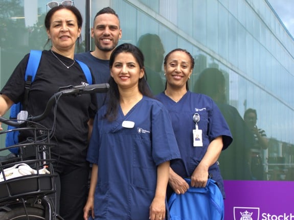 Fyra personer som jobbar inom hemtjänsten i Skärholmen. De poserar framför kameran med en cykel och en del av Stockholm stads logo syns i vänstra nedre hörnet i bilden