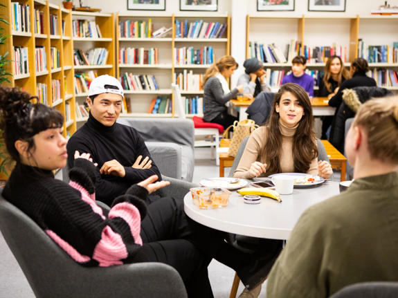 Fyra deltagare sitter runt ett bord i skolans kombinerade bibliotek och fikarum. En av dem äter lunch och de andra sitter vid bordet och pratar