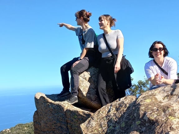Tre tjejer står på en klippa och tittar ut mot havet. Himlen är klarblå och den ena tjejen pekar ut mot horisonten.