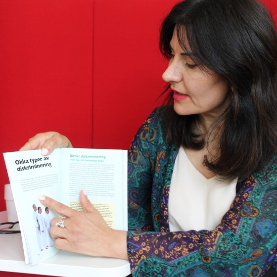 Läraren Meral Onmaz intervjuas om kursen Agera utan att diskriminera. Meral sitter med en röd bakgrund och visar på ett projektarbete tidigare deltagare har gjort