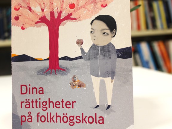 Vägg i skolans reception med en vepa med Röda Korsets folkhögskolas värdeord. Till höger om vepan en anslagstavla med olika informationslappar uppsatta. Under anslagstavlan står en rödmålad parkbänk. Högst upp på ryggen står skrivet