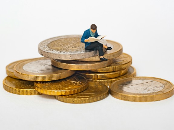 På en trave med mynt sitter en liten docka med korslagda ben och läser i en bok