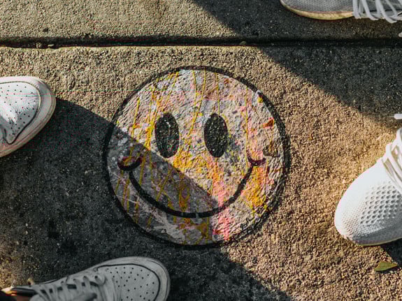 En smiley ritad med kritor fotad uppifrån på en gata. På var sida om smileyn ser man fötterna på två personer. De har sneakers på sig