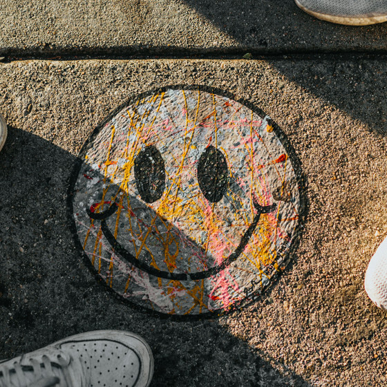 En smiley ritad på en trottoar med krita i olika färger. Bilden är tagen uppifrån och man ser också två personers fötter stå bredvid smileyn. De har sneakers på sig