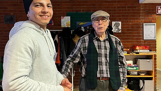 En ung manlig deltagare från folkhögskolan i svart mössa står bredvid en äldre man med rullator och keps. De är på seniorcaféet i Skärholmen.