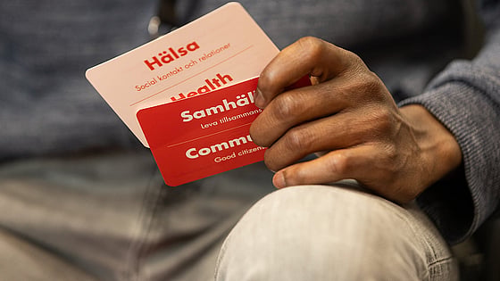 Händerna på en person syns i bild. Personen håller i två kort. Ett står det hälsa på, det andra står det samhälle på.