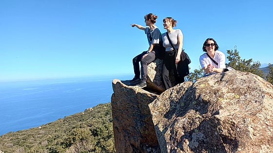 Tre tjejer står på en klippa vid medelhavet. Himlen är klarblå och en av tjejerna pekar ut mot havet.
