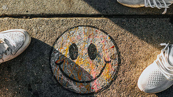 En smiley ritad på en trottoar med krita i olika färger. Bilden är tagen uppifrån och man ser också två personers fötter stå bredvid smileyn. De har sneakers på sig