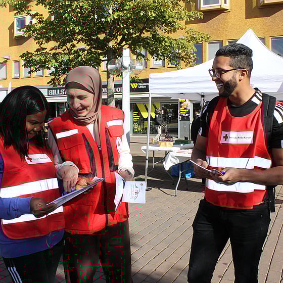 Tre deltagare från Röda Korsets folkhögskola delar ut material om krisberedskap på torget i Skärholmen. De har rödakorsvästar och är glada.