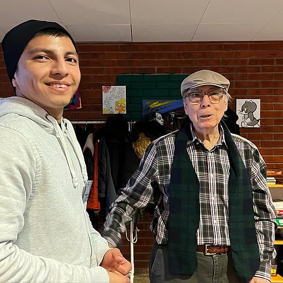 En ung manlig deltagare från folkhögskolan i svart mössa står bredvid en äldre man med rullator och keps. De är på seniorcaféet i Skärholmen.