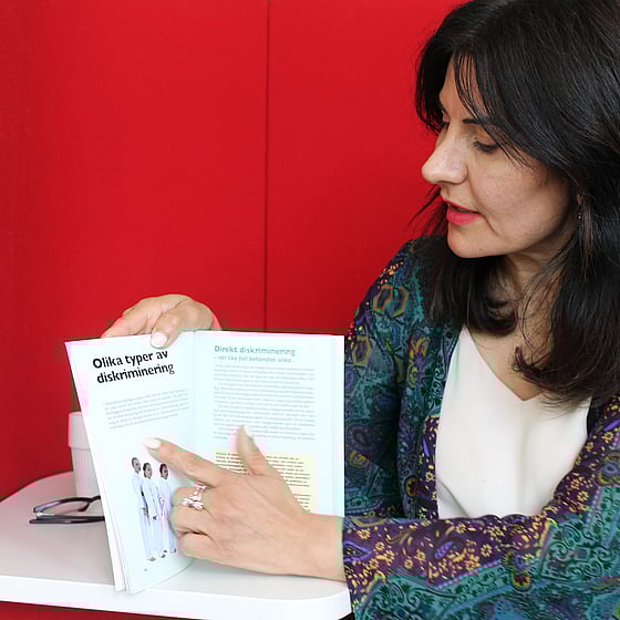 Läraren Meral Onmaz intervjuas om kursen Agera utan att diskriminera. Meral sitter med en röd bakgrund och visar på ett projektarbete tidigare deltagare har gjort
