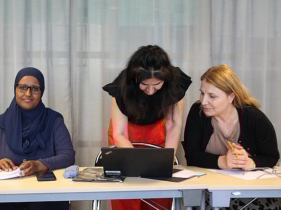 Två kvinnliga kursdeltagare jobbar med ett skolarbete och en lärare instruerar