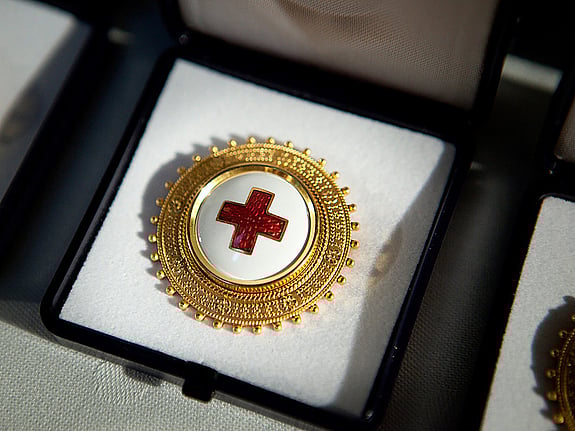 En brosch med Röda Korsets symbol i mitten på vit botten och en kant av guld. Broschen ligger i ett fodral på ett bord.