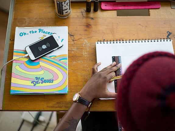 En kille i röd mössa syns bakifrån när han pluggar matte. Närbild på baksida huvud, arm med klocka och skrivbord med telefon och skrivböcker