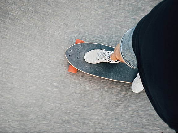 En skateboard sedd uppifrån på asfalt. Man ser fötterna på personen som åker skateboard.