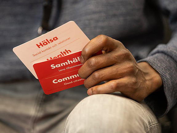 Man ser händerna på en person som sitter ned med grå tröja och vita byxor. Personen håller i två samtalskort. Det ena står det hälsa på. Det andra står det samhälle på