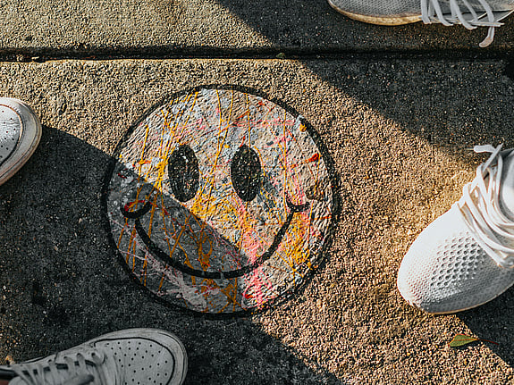 En smiley ritad med kritor fotad uppifrån på en gata. På var sida om smileyn ser man fötterna på två personer. De har sneakers på sig