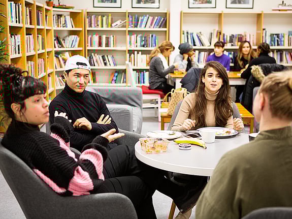 Fyra deltagare sitter runt ett bord i skolans kombinerade bibliotek och fikarum. En av dem äter lunch och de andra sitter vid bordet och pratar