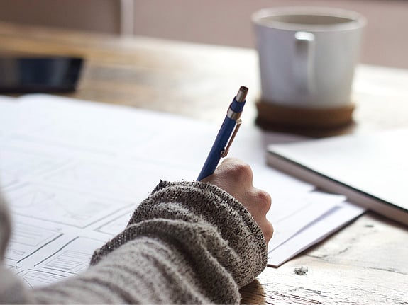 närbild hand som skriver. i bakgrunden står en mugg