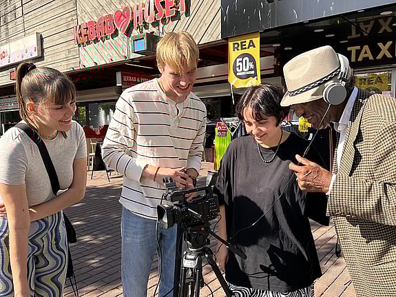 Fyra personer står och tittar in i en kamera på stativ som står framför dem. De gör en filminspelning i Skärholmens centrum