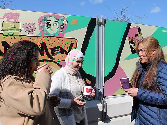 Tre kvinnor står framför en grafittivägg och dricker kaffe ur pappersmuggar