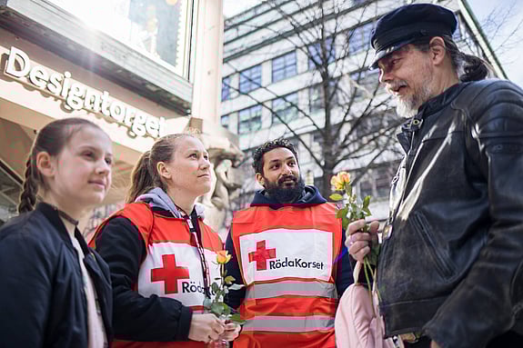 Fyra personer står på en gata i en stad. Till vänster en ung flicka. I mitten en kvinna och en man i Röda Korsvästar och till höger en lång man med keps. Han håller en blomma i handen. Personerna i mitten är krisstödjare från Röda Korset