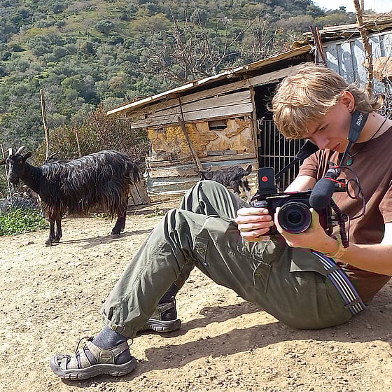 En ung man sitter på marken och ställer in sin kamera. Bakom honom ser man ett skjul och en svart get. I bakgrunden syns ett berg med växtlighet