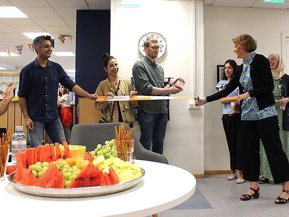 Rektor klipper bandet vid en releasefest för de nya Pratsamtkorten. Två personer håller i bandet och i fronten står en fruktskål med vattenmelon