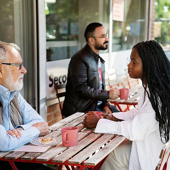 Fyra personer sitter utomhus vid cafébord. De för samtal två och två. Det är både äldre och yngre personer.
