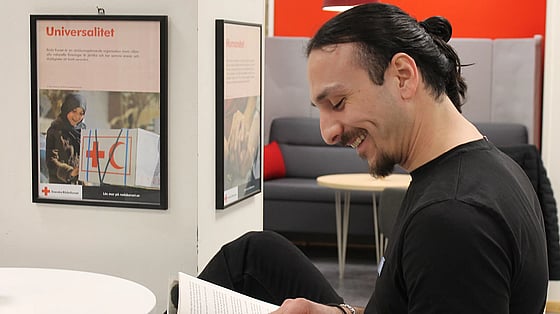 En ung man sitter och tittar ner i sitt block. Han ser glad ut. Bakom honom syns tavlor som beskriver Röda Korsets grundprinciper