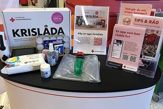 På ett bord står olika produkter som kan ingå i en krislåda. Det är värmeljus, tändstickor, en hopfällbar vattendunk, våtservetter och en kortlek. Det finns också broschyrer och informationsmaterial från Röda Korset om krisberedskap.