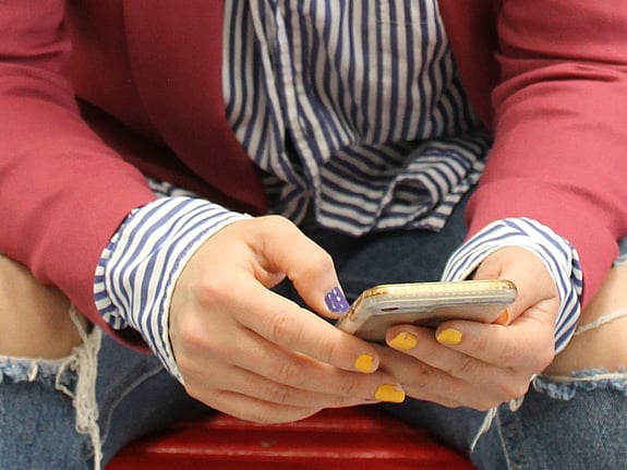 Man ser bara händer och knän på personen på bilden. I händerna håller hen en mobiltelefon. Hen ha röd kavaj, randig skjorta och jeans som är trasiga över knäna.