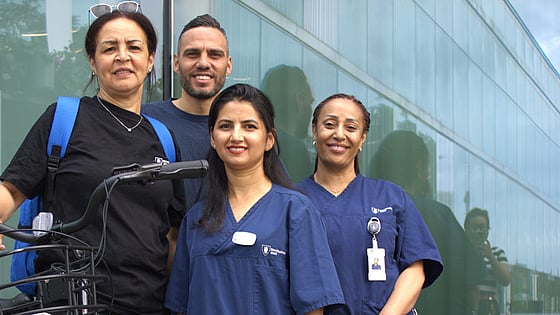 Fyra personer står tillsammans framför en vägg. De har alla hemtjänstkläder på sig. Det är tre kvinnor och en man. En av kvinnorna håller i en cykel