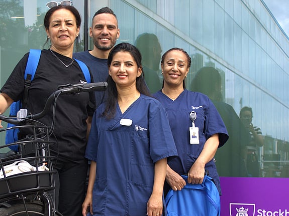 Fyra personer som jobbar inom hemtjänsten i Skärholmen. De poserar framför kameran med en cykel och en del av Stockholm stads logo syns i vänstra nedre hörnet i bilden