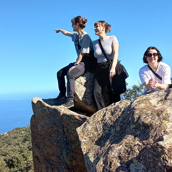 Tre tjejer som går en dokumentärfilmskurs tittar ut över medelhavet från en klippa i Sicilien. Solen är skarp och havet är väldigt blått. En tjej pekar ut mot horisonten.