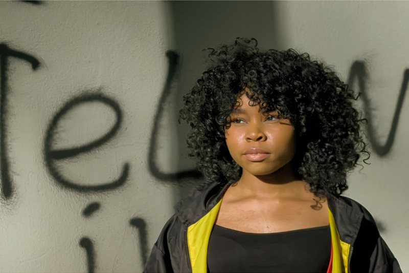 En mörkhyad tjej med bestämd och fundersam blick står fram för en vit vägg med svart sprejad text.