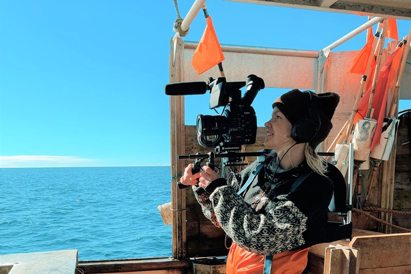 En kvinnlig dokumentärfilmare i islandströja, svart stickad mössa och orangea fiskebyxor står på en fiskebåt och filmar med en stor svart filmkamera. Himlen är klarblå, solen lyser och även havet är härligt blått.