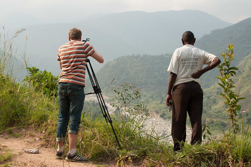 En dokumentärfilmare står och filmar en grön dal mellan bergen i Uganda. En lokalinvånare står bredvid och blickar ut över dalen.