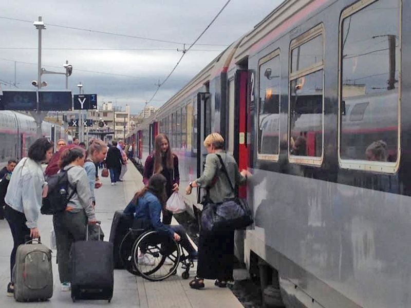 Deltagare står på en tågstation och ska precis gå på ett tåg. De har sitt bagage i händerna. De hjälper en av dem som sitter i rullstol att komma på först. 