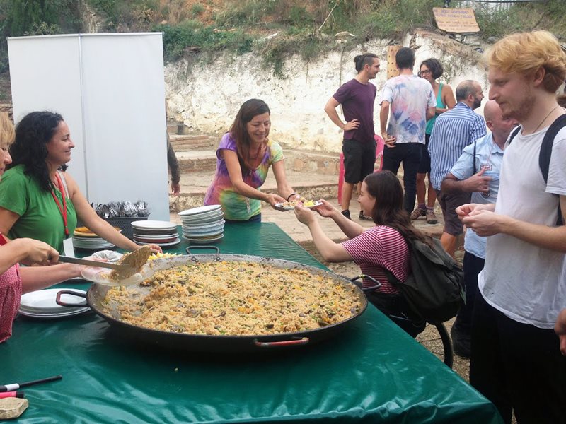 Två kvinnor står till vänster om ett bord och serverar paella från ett stort fat. På högra sidan står några deltagare och väntar på att få sin mat. På bordet ligger en grön plastduk under paellapannan.