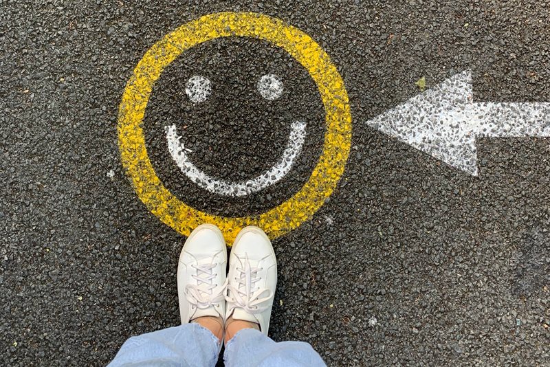En  glad smiley målad på asfalt. En vit pil som pekar på smileyn. Nedre delen av ett par ben med ljusa jeans och vita gympaskor står i riktning mot smileyn. 