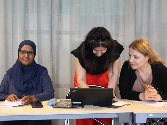 Två kvinnliga kursdeltagare jobbar med ett skolarbete och en lärare instruerar