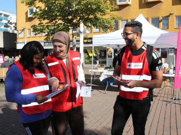 Tre deltagare från Röda Korsets folkhögskola står på torget i Skärholmen och delar ut information om krisberedskap. De är glada.