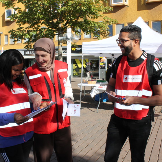 Tre deltagare från Röda Korsets folkhögskola delar ut material om krisberedskap på torget i Skärholmen. De har rödakorsvästar och är glada.