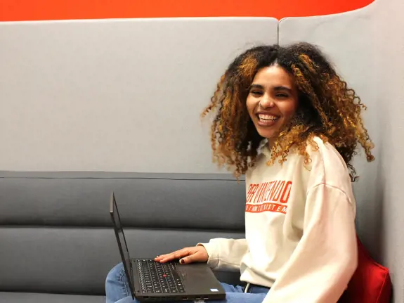 Ung flicka sitter i soffa med dator i knät. Hon skrattar in i kameran