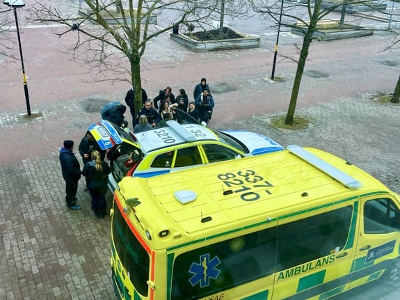 Deltagare från Röda Korsets folkhögskola står samlade utanför skolan vid en polisbil och en ambulans. Två poliser visar bilen och berättar om yrket. Det är den återkommande Blåljusdagen på Röda Korsets folkhögskola