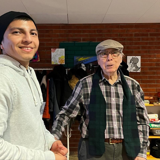 En ung manlig deltagare från folkhögskolan i svart mössa står bredvid en äldre man med rullator och keps. De är på seniorcaféet i Skärholmen.