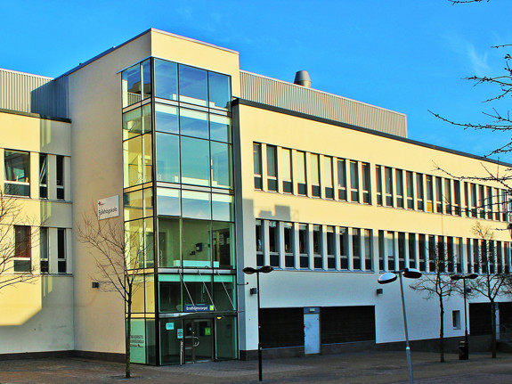 En vit byggnad i tre våningar med ett stort glasat trapphus och många fönster i rader till höger om trapphuset. Ovanför huset är himlen klarblå.